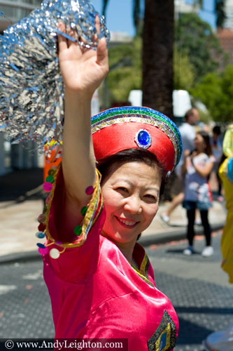 A smiling red costumed Chinese woman looks at the camera and raises her hand into the air as she parades along James Street. Australian Mulan Arts & Culture Association (AMACA), WA Chinese Woman's Federation (WACWF) Chinese New Year 2013, Northbridge, Perth, Australia