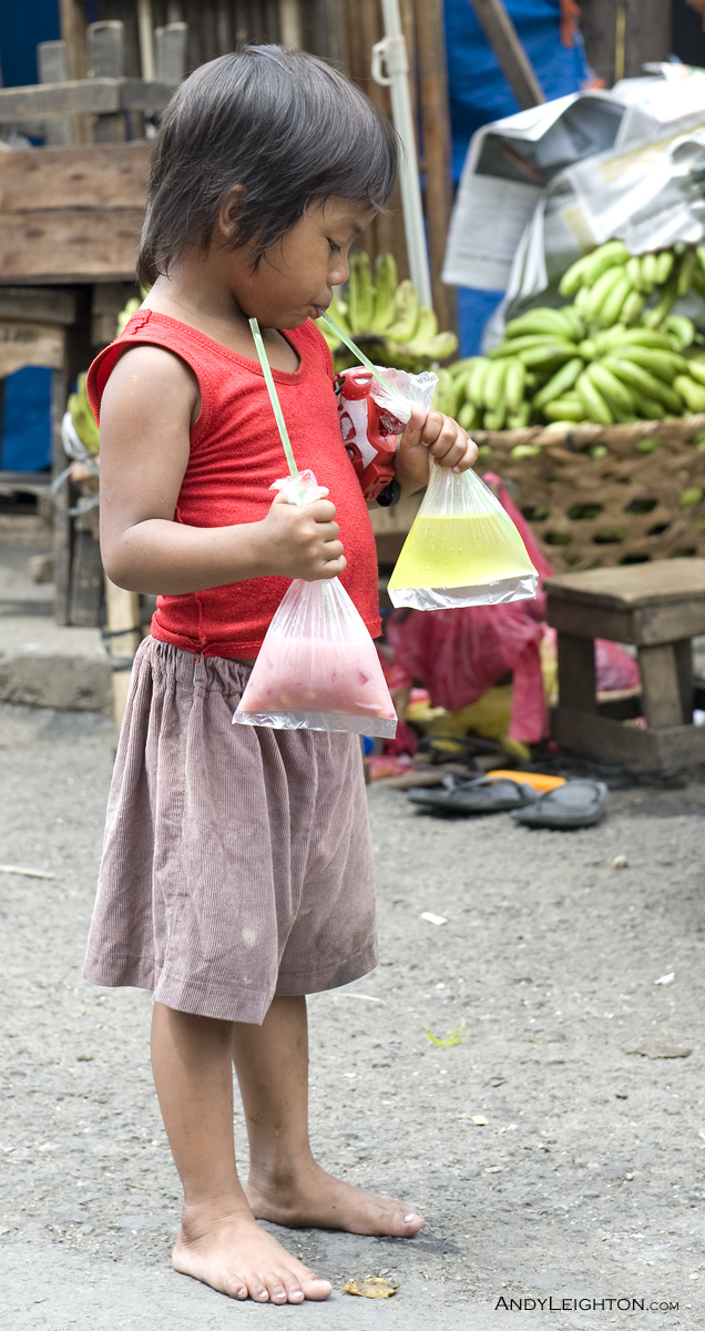 A young boy drinks fruit juice through a straw from one of two plastic bags he is holding, Carbon Market, Philippines